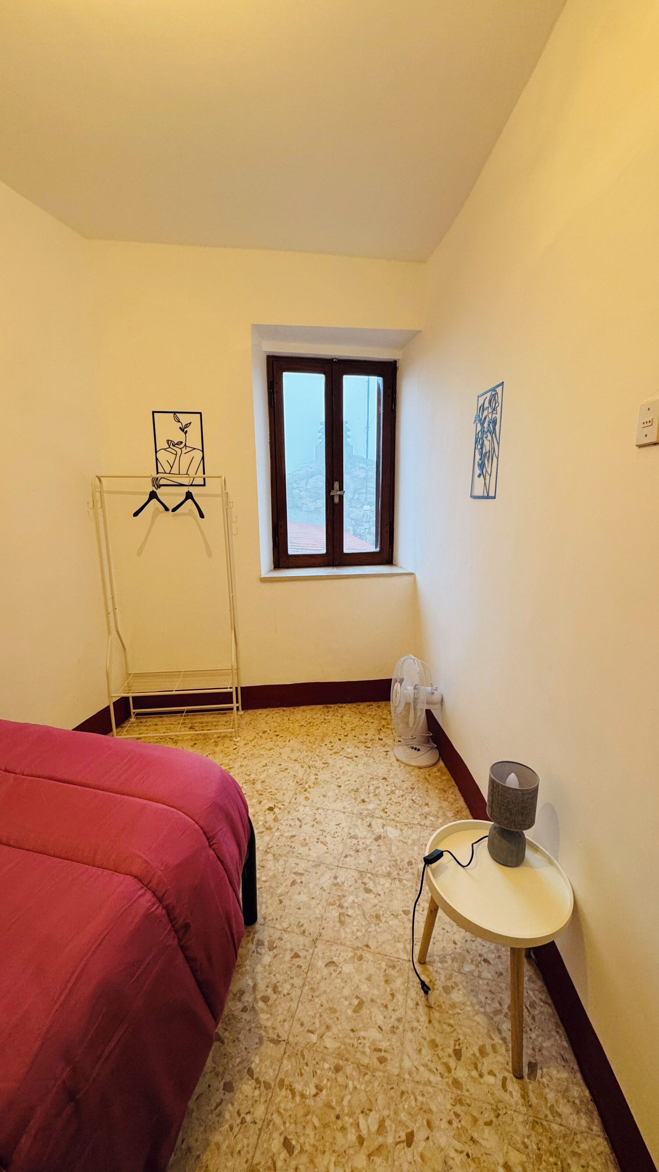 A room with a bed, a window, a fan, and a small table.