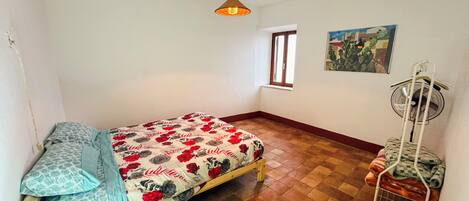 A bedroom with a bed, a window, a painting, a fan, and a wooden floor.
