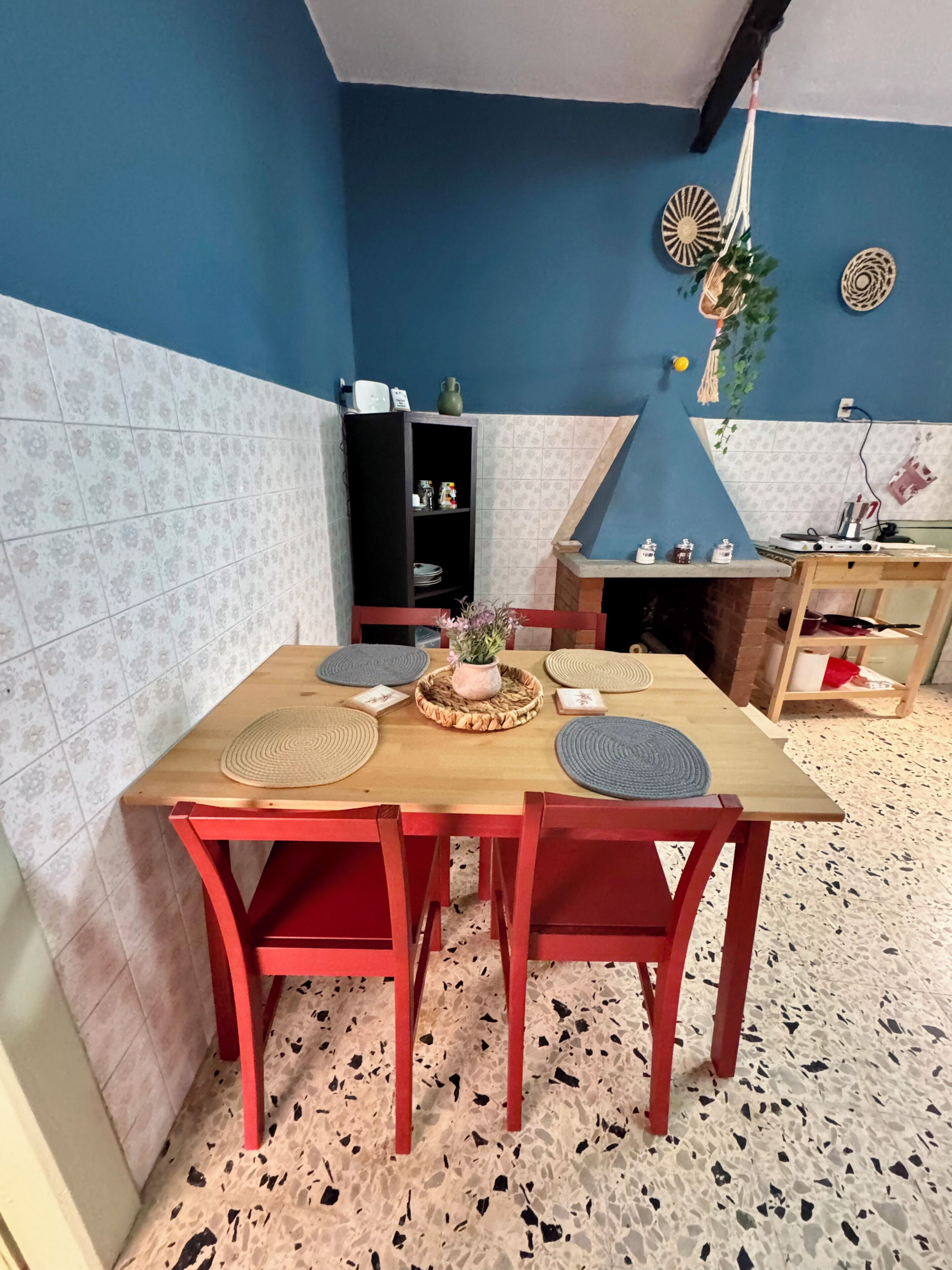 A dining area with a wooden table and red chairs, a black shelf with kitchen items, and a blue wall with decorative plates.