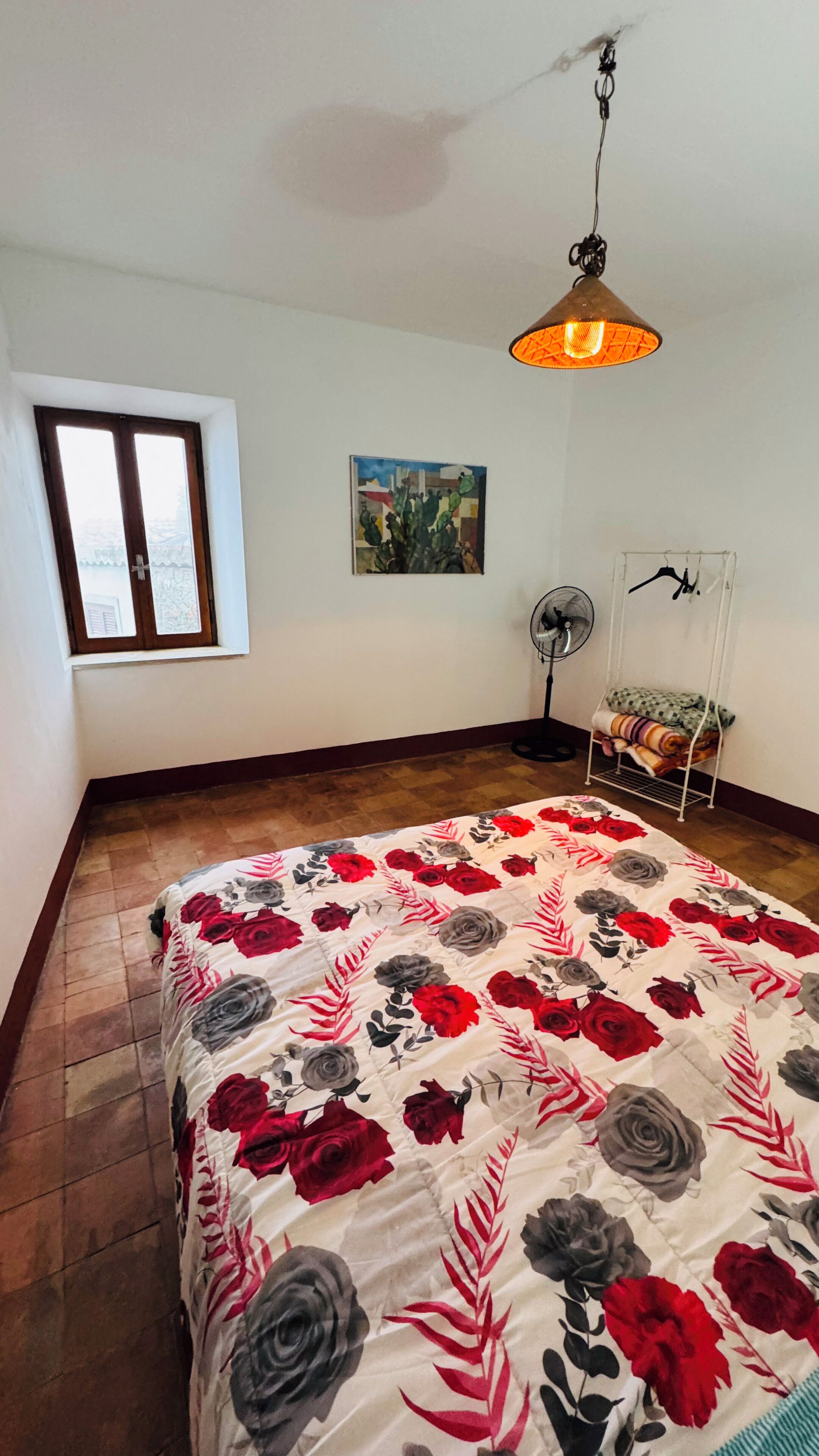 A bedroom with a bed, a window, a painting, a standing fan, and a heater.
