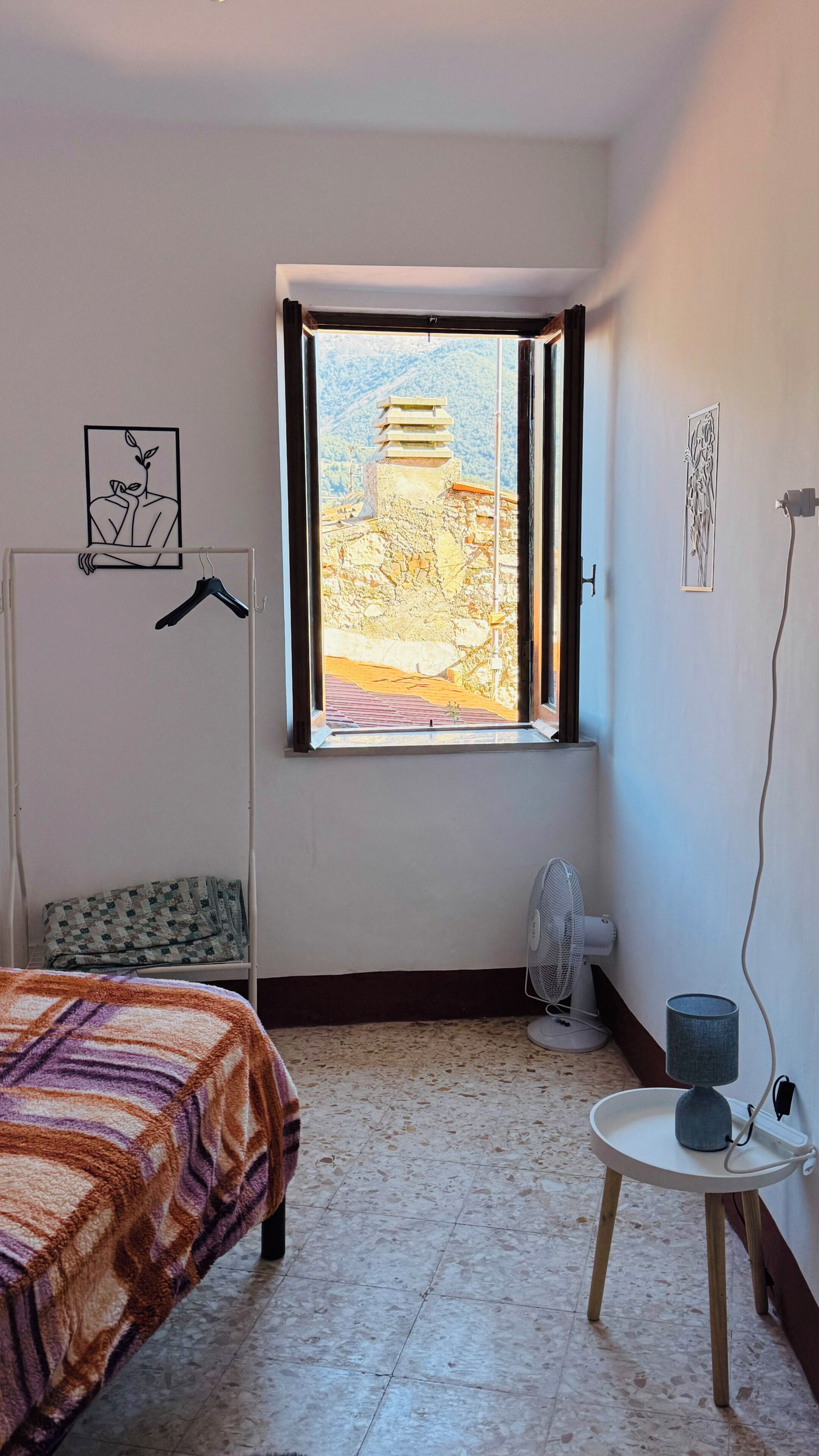A bedroom with a bed, a window with a view of a building, a radiator, and a small table with a lamp.