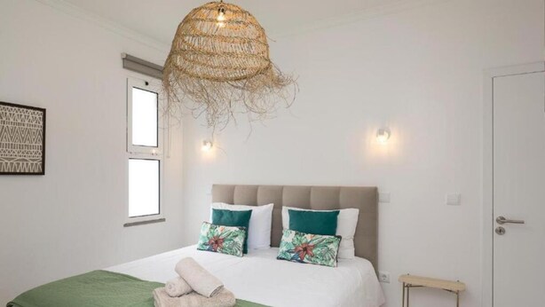 A neatly made bed with a green blanket and two pillows, a woven ceiling light fixture, and a small wooden side table.