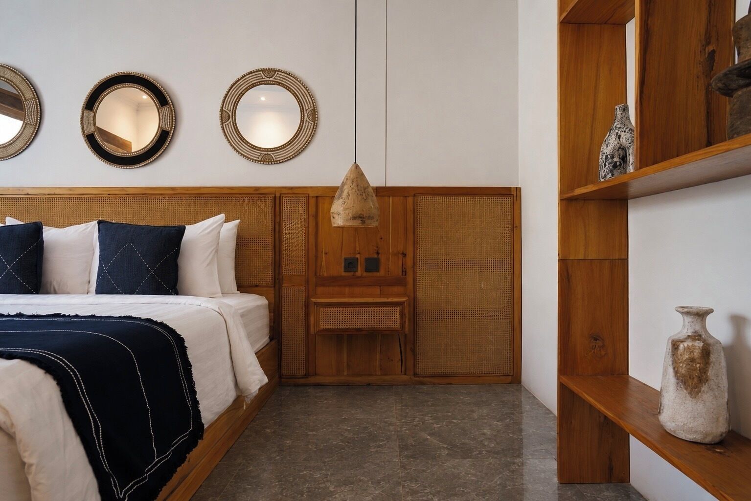 A bedroom with a bed, three wall-mounted mirrors, a woven headboard, a hanging lamp, and a shelf with a vase.