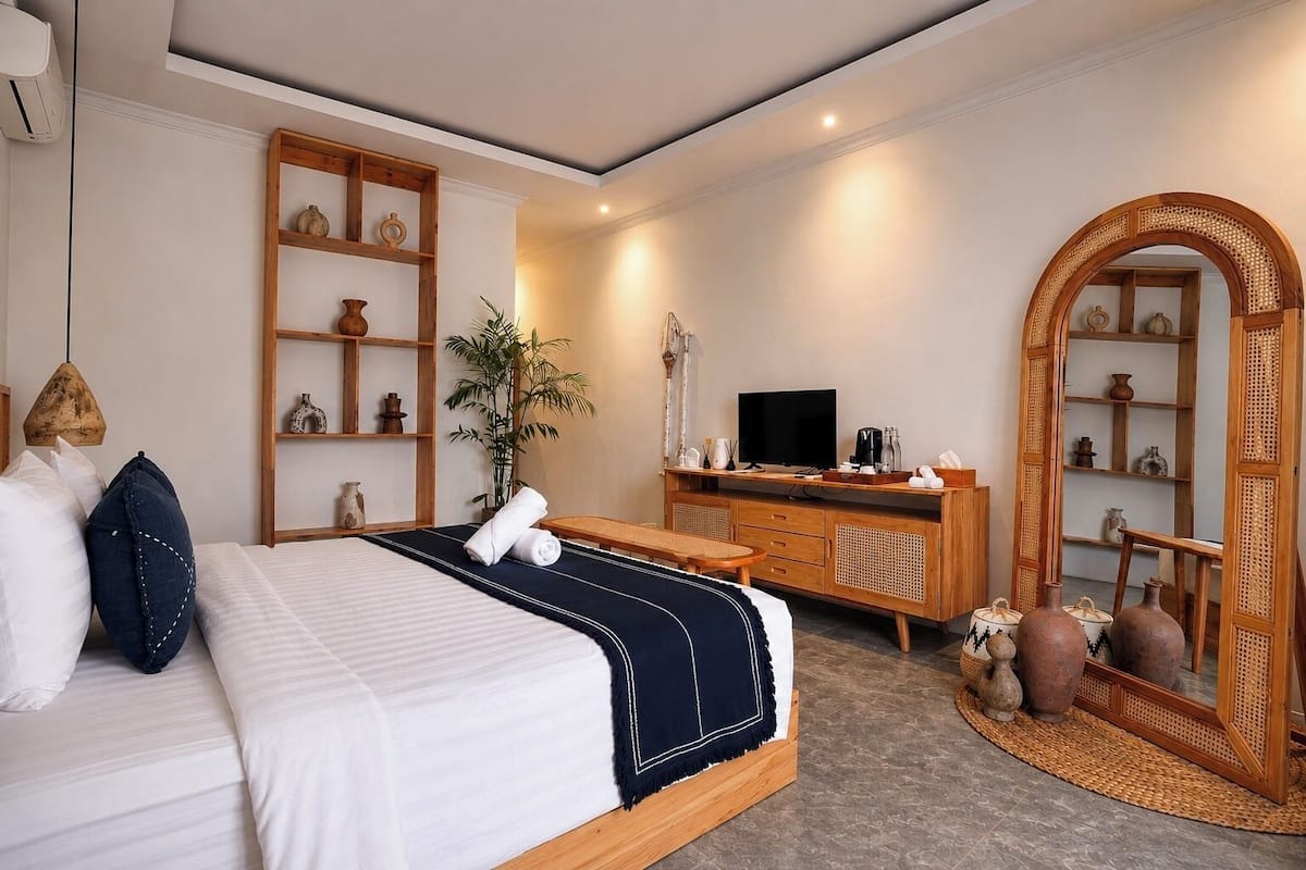 A hotel room with a bed, a desk with a flat-screen TV, a mirror, and shelves with decorative items.
