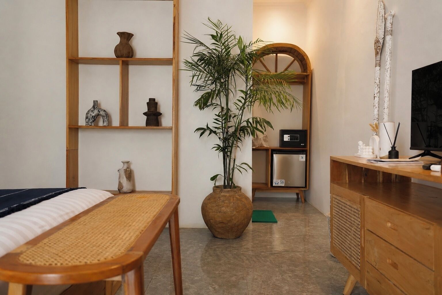 A room with a bed, a wooden desk, a large potted plant, a small refrigerator, and a television.