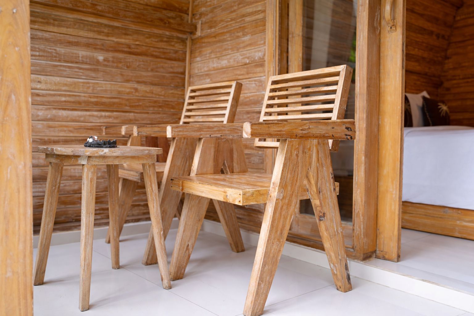 A wooden cabin interior with two wooden chairs and a small table, a bed with a white mattress, and a wooden floor.