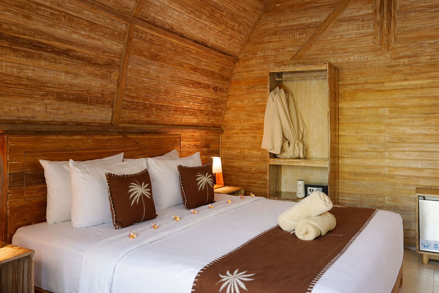 A wooden-paneled room with a bed, pillows, a nightstand, a robe, and a small table with a lamp.