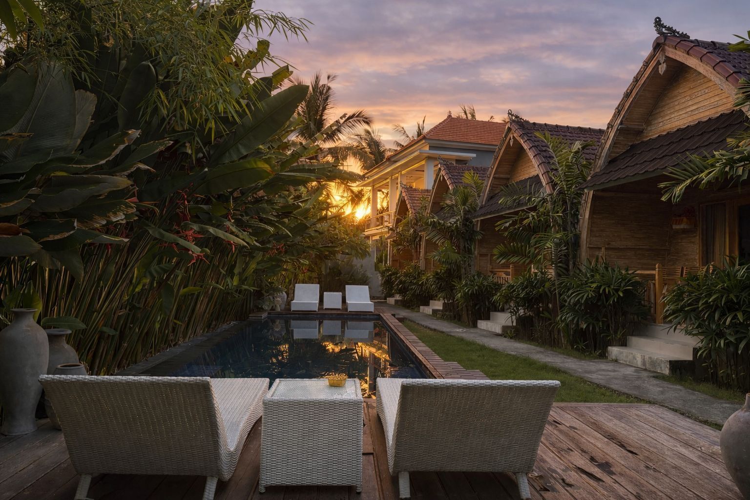 A pool area with lounge chairs, surrounded by tropical plants and wooden buildings.