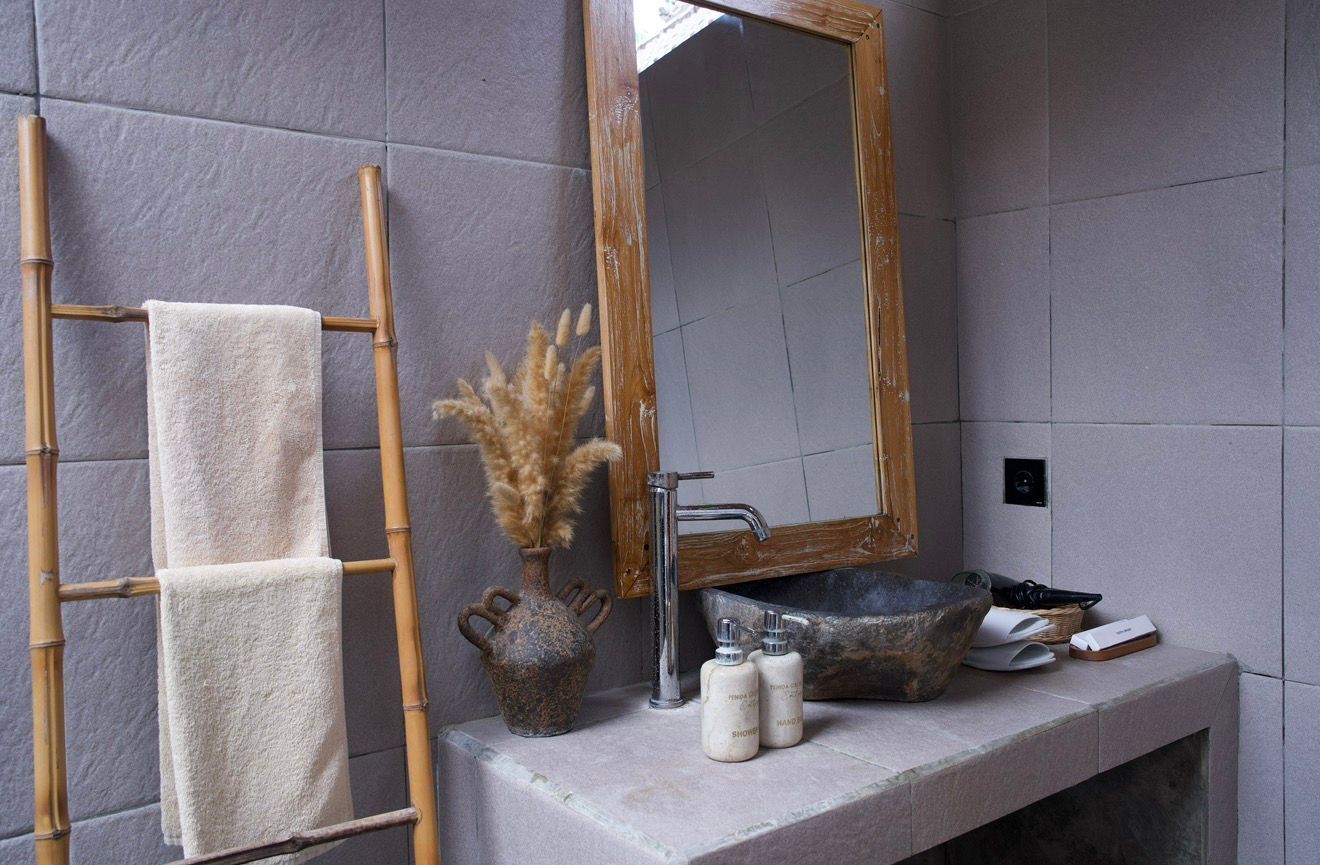 A bathroom with a bamboo towel rack, a mirror, a vase with dried flowers, a sink with a stone basin, and toiletries.