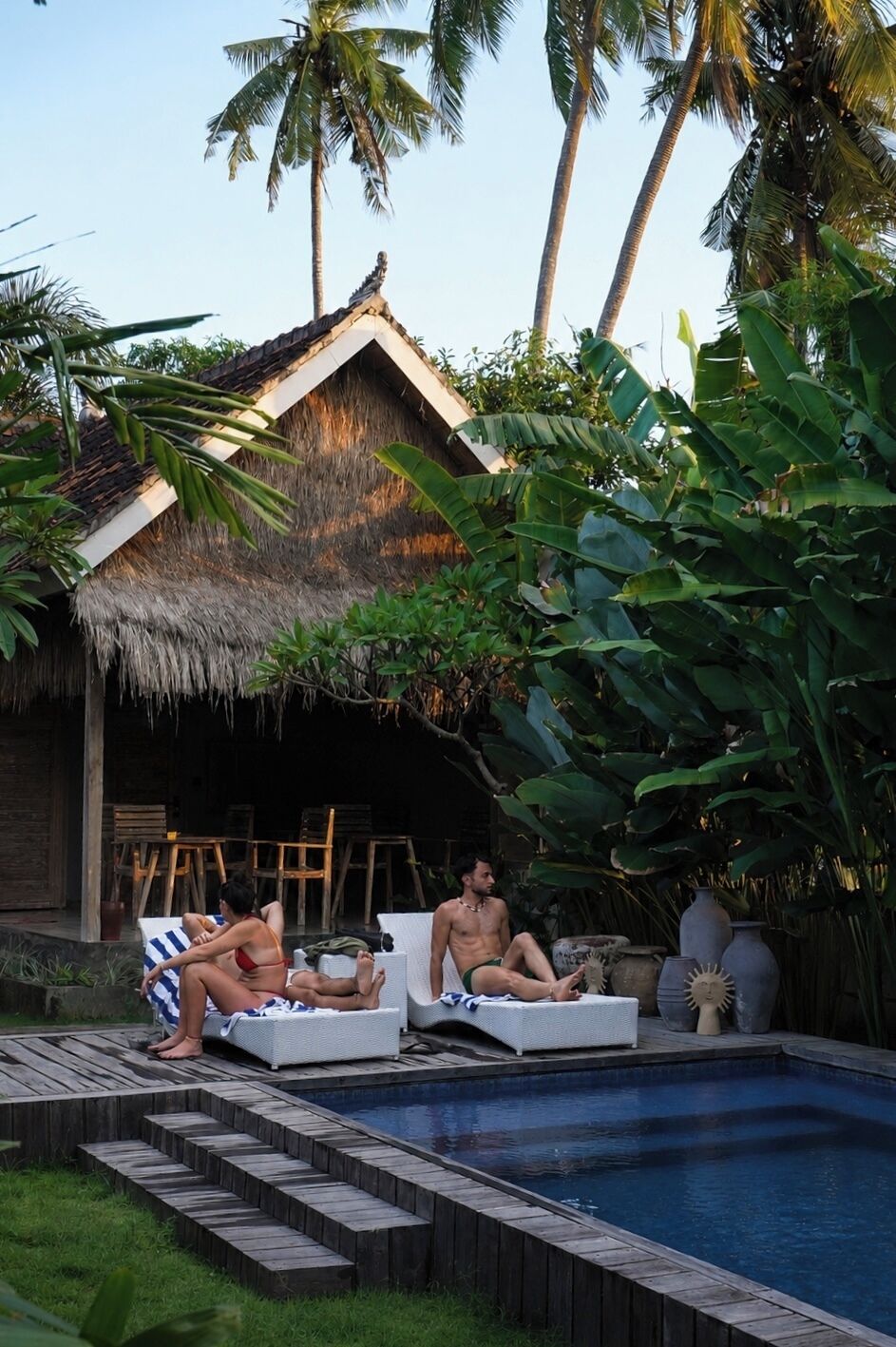 A poolside area with lounge chairs, a thatched-roof structure, and tropical vegetation.