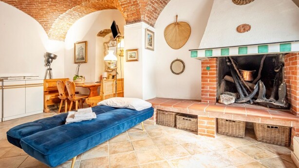 Een kamer met een haard van baksteen, een blauwe bank, een eethoek met tafel en stoelen, en een keuken met kastjes.