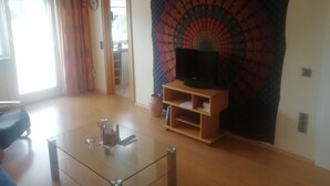 Een woonkamer met een glazen salontafel, een flatscreen televisie op een houten standaard, een wandtapijt aan de muur en een deur naar een andere kamer.