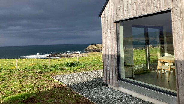 A modern wooden building with a large glass window, a gravel pathway, and a view of the ocean. - Wow sea-view! walks, cliffs, wildlife - spacious contemporary home (Portree)