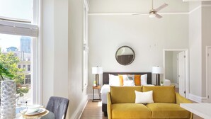 A modern hotel room with a large window, a sofa, a bed, and a ceiling fan.