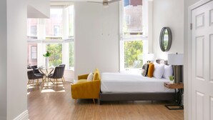 A modern bedroom with a large bed, a dining area with chairs, and a view of the outside.