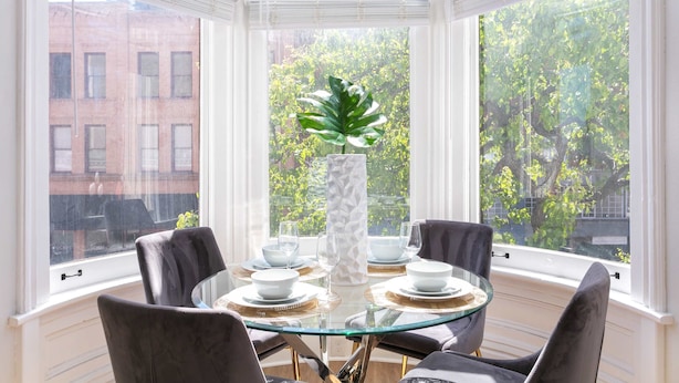 A dining area with a glass table surrounded by chairs, a tall white vase, and a view of trees and buildings through large windows.