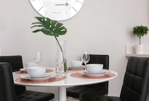 Une table de salle à manger ronde, surmontée d’assiettes, de bols et de verres à vin blancs, est entourée de chaises noires à revêtement en tissu. Un grand miroir est accroché au mur, au-dessus de la table.
