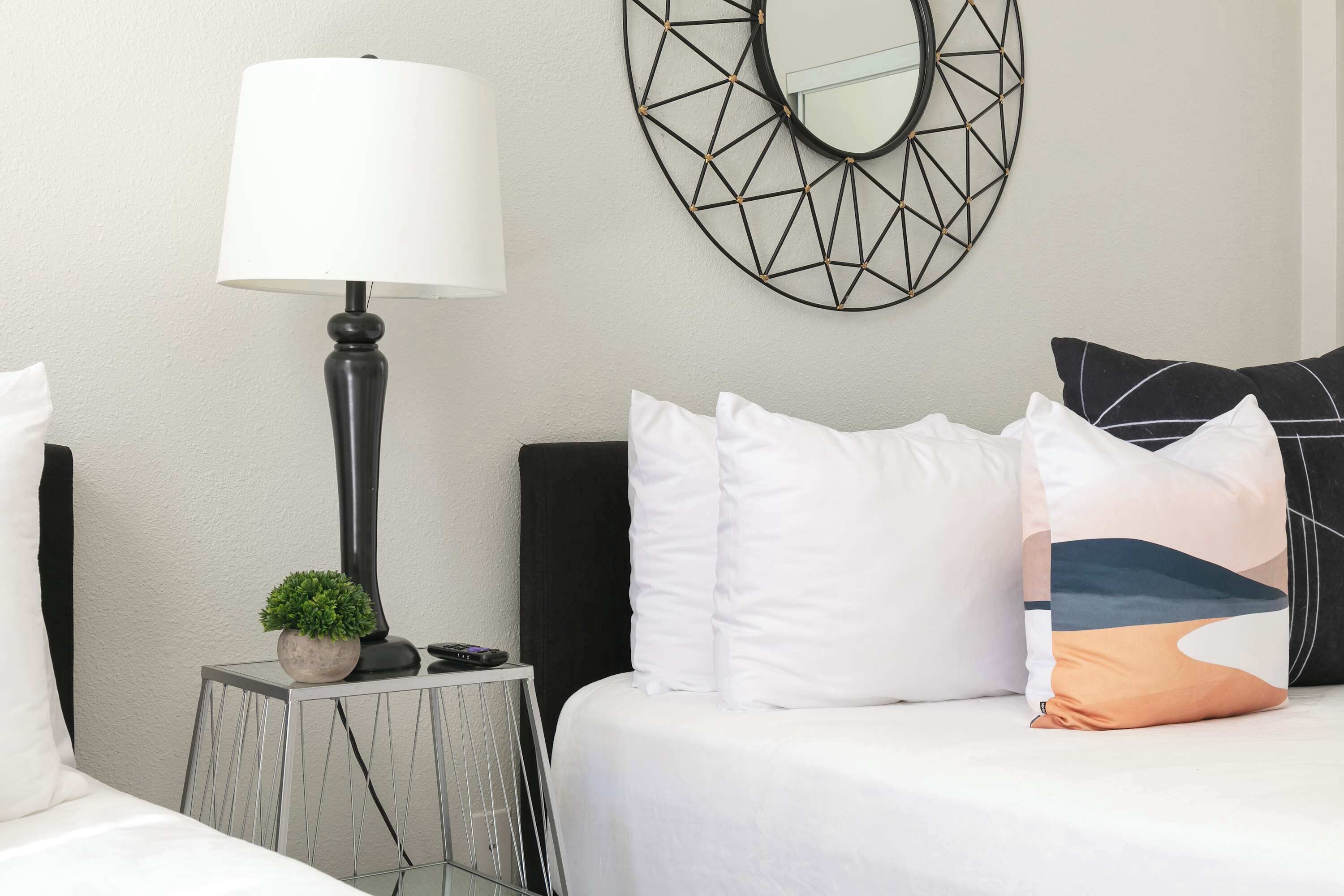 A neatly arranged hotel room with a bed, a bedside table with a lamp, a small plant, and a decorative wall piece.