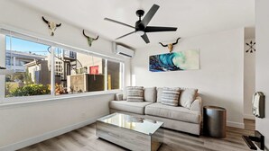 Una sala de estar moderna con una mesa de centro de vidrio, un sofá color beige, un ventilador de techo y un ventanal que ofrece vistas al exterior.