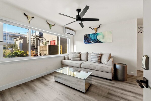 A modern living room with a glass coffee table, a beige sofa, a ceiling fan, and a large window offering a view of the outside.