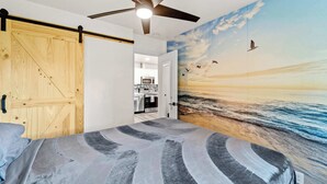Un dormitorio con una cama, un ventilador de techo, una puerta de madera y un mural con una escena de playa.