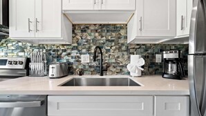 A modern kitchen with white cabinets, a stainless steel sink, a toaster, a coffee maker, and a tiled backsplash. - 2BR Urban Retreat w/ Rooftop Lounge (San Diego)