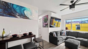 A modern living room with a dining area, grey sofas, a ceiling fan, and a large window with a view of a yellow building. - 2BR Urban Retreat w/ Rooftop Lounge (San Diego)