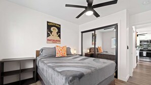 A modern bedroom with a large bed, a ceiling fan, a wardrobe, and a kitchen visible through an open door. - 2BR Urban Retreat w/ Rooftop Lounge (San Diego)