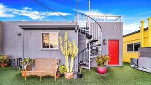 A modern building with a grey exterior, a red door, and a balcony. There are potted plants and a wicker chair outside.