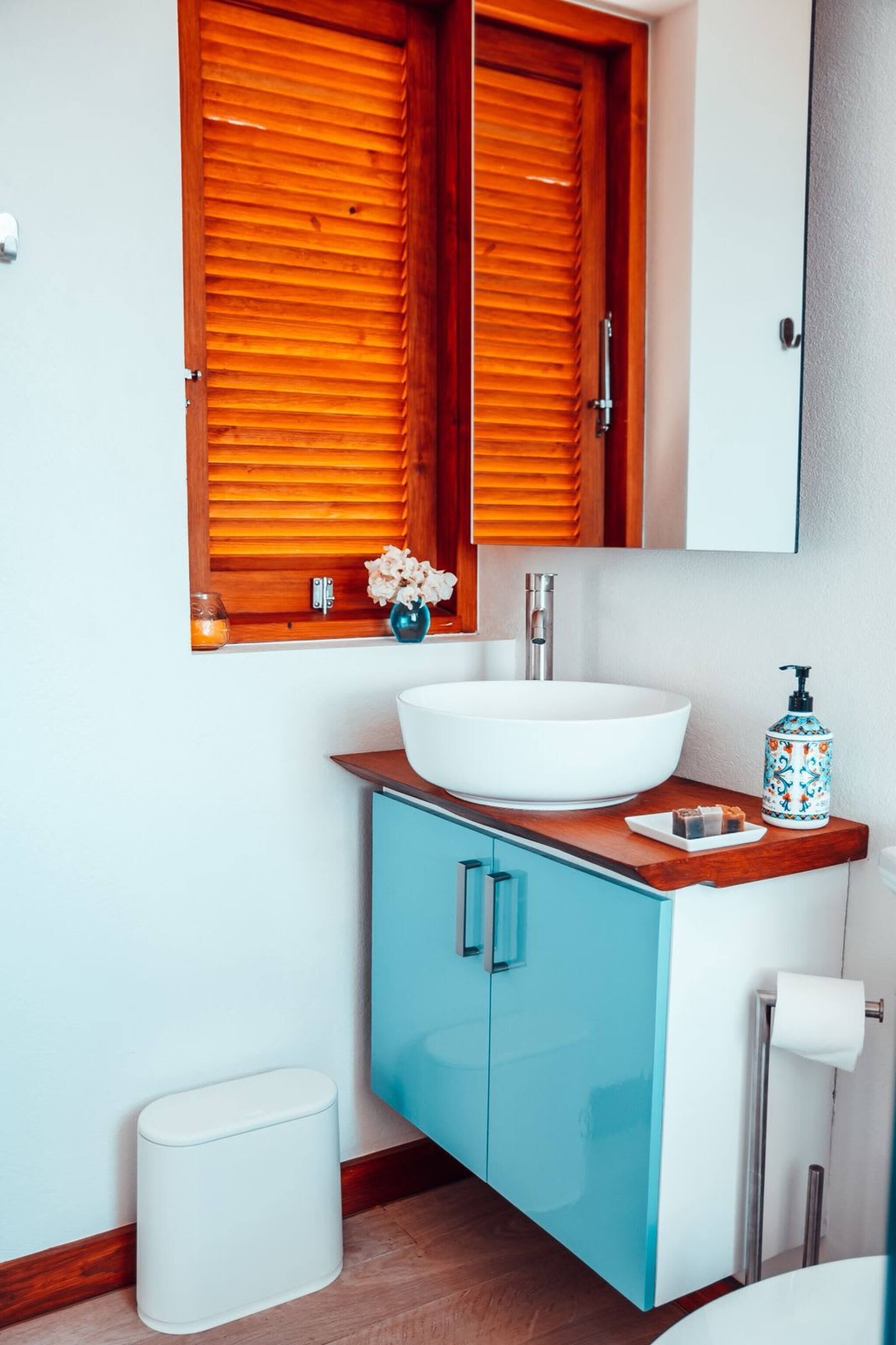 Un baño con lavamanos blanco, muebles azules, persianas de madera, un jarrón pequeño con flores y un cubo de basura blanco.