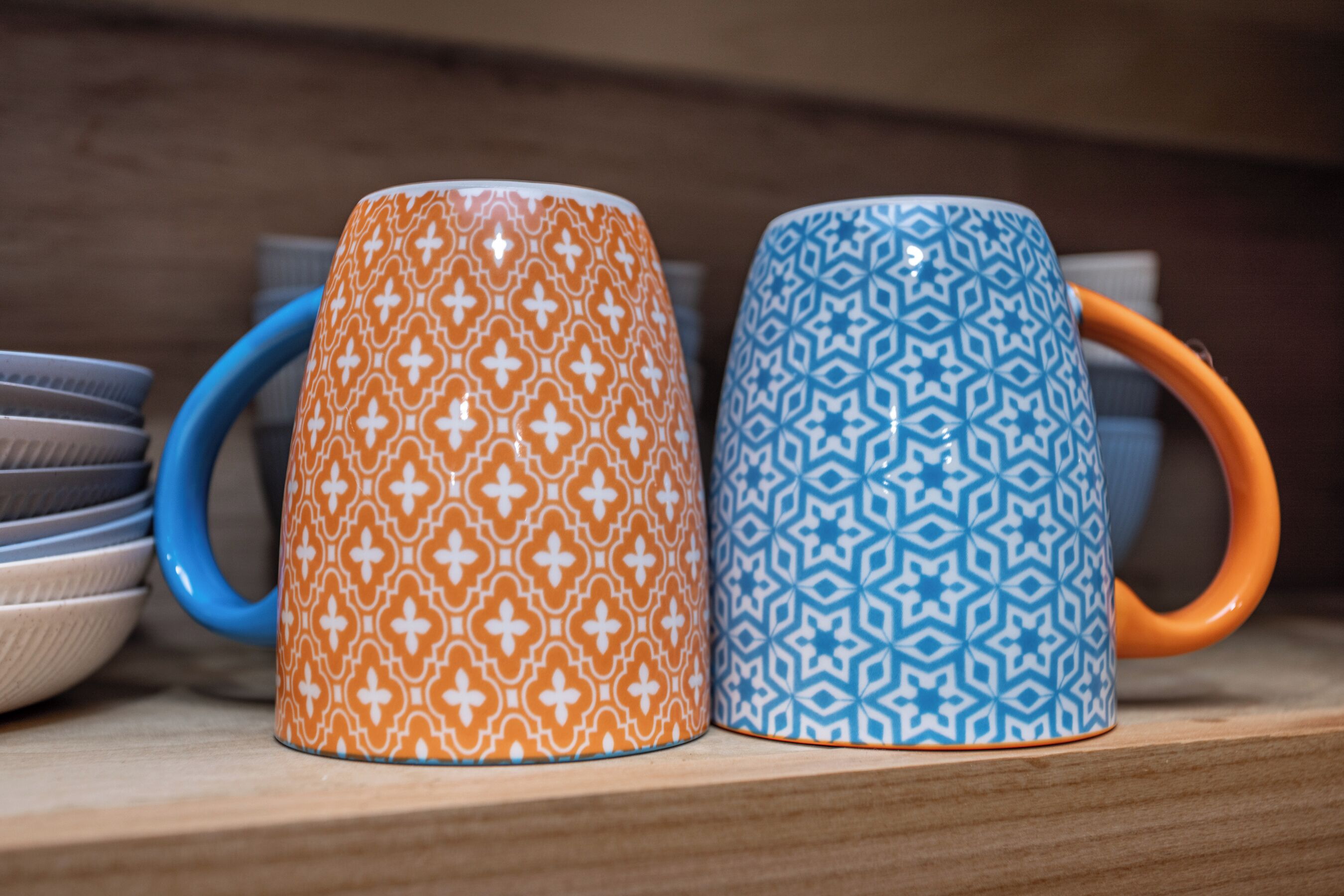 Two patterned mugs with handles, placed on a shelf with other items.