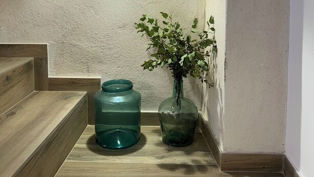 Eine Ecke mit einer Holztreppe, einem grünen Wasserspender und einer Vase mit grünen Blättern.