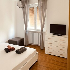 Ein Schlafzimmer mit einem Bett, einem Fernseher auf einer Kommode, einem Heizkörper und einem Fenster mit Vorhängen.