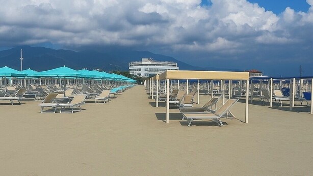 A beach with rows of sun loungers under blue and beige umbrellas, a large building in the background, and mountains in the distance. - Detached house in the center of Tonfano • 100 m from the sea • 2 parking spaces (Marina di Pietrasanta)