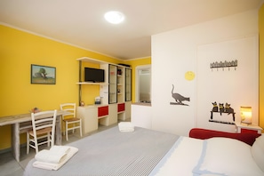 Una habitación con cama, escritorio, silla, armario, televisor y un mural de un gato en la pared.