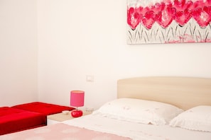 Un dormitorio con cama, una mesita de noche, una lámpara roja y un cuadro de flores.