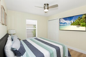 A bedroom with a bed, a ceiling fan, a window with blinds, a wardrobe, and a TV displaying a beach scene.