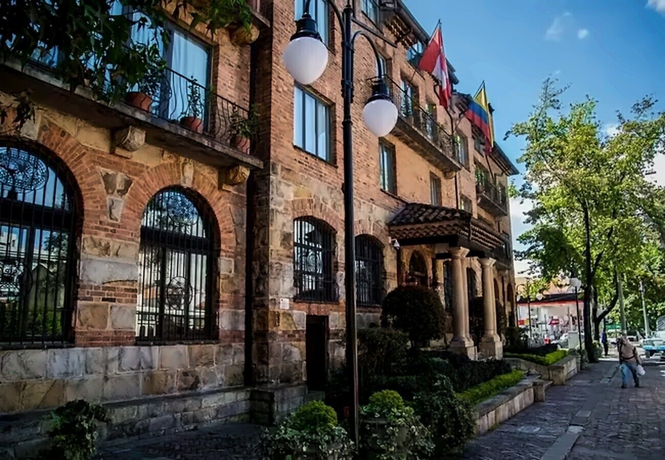 A historic building with arched windows and a balcony, surrounded by greenery and a paved walkway.