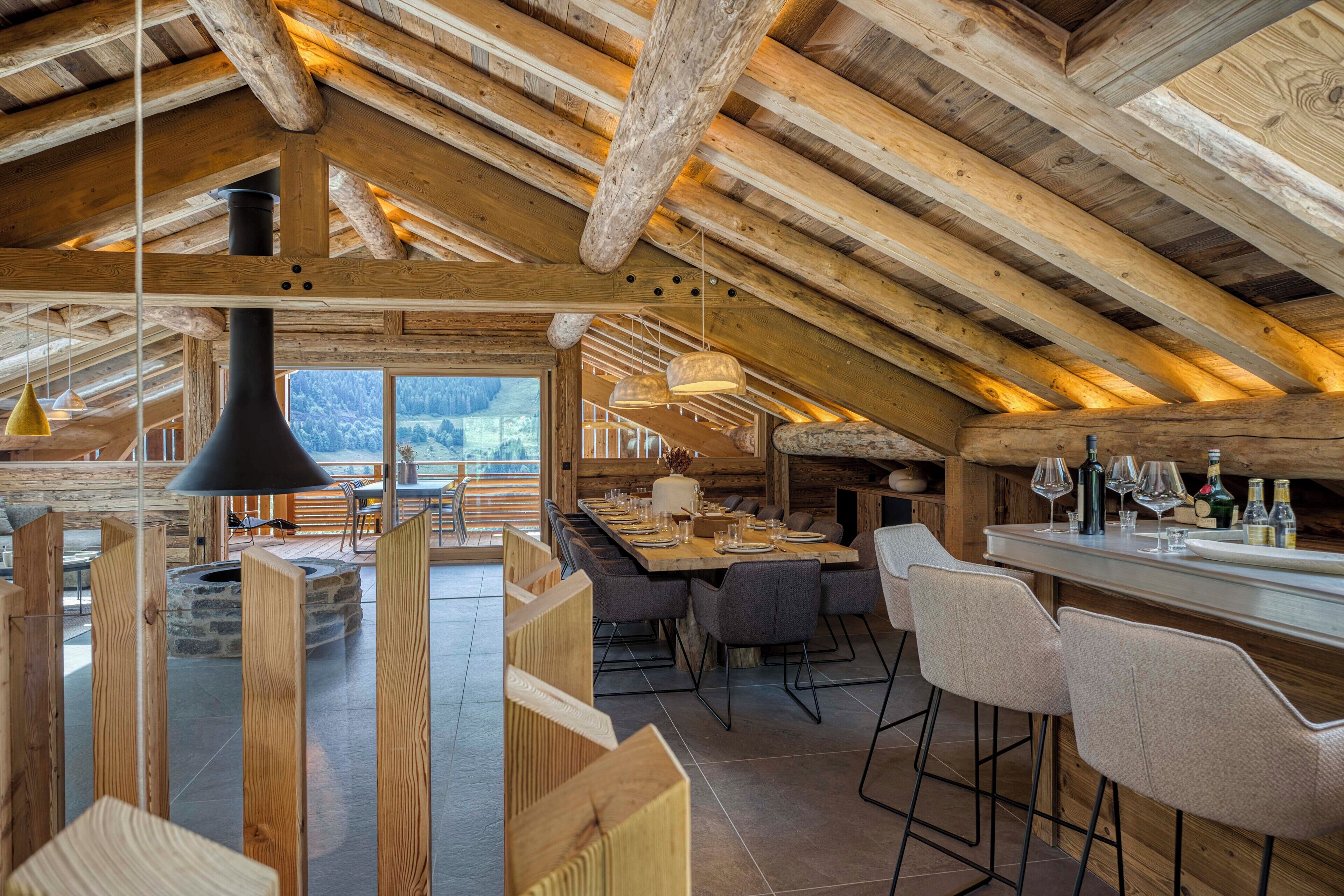 Un intérieur rustique en bois, avec un plafond haut, un coin repas doté d’une longue table et de chaises, et un bar avec des bouteilles et des verres.