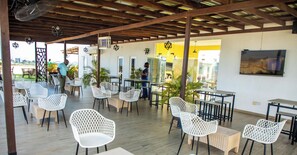 A covered outdoor seating area with white wicker furniture and a wooden floor, featuring a TV mounted on the wall.