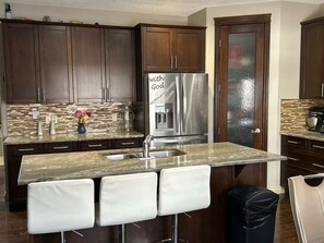 A modern kitchen with dark wood cabinets, a stainless steel refrigerator, and a granite countertop. - Spacious 4BR Retreat- Perfect for Families & Groups! (Airdrie)