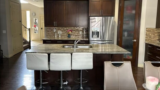 A modern kitchen with dark wood cabinets, a stainless steel refrigerator, a marble countertop, and three bar stools. - Spacious 4BR Retreat- Perfect for Families & Groups! (Airdrie)