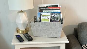 A white nightstand with a lamp, remote controls, a basket of books, a red notebook, and a wicker basket.