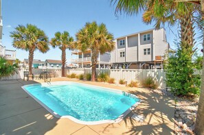 Ein Schwimmbecken mit Holzterrasse, umgeben von Palmen und einem weißen Zaun.