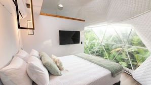 Une chambre avec un grand lit, du linge de lit blanc, un téléviseur fixé au mur et une vue sur la verdure par une grande fenêtre.