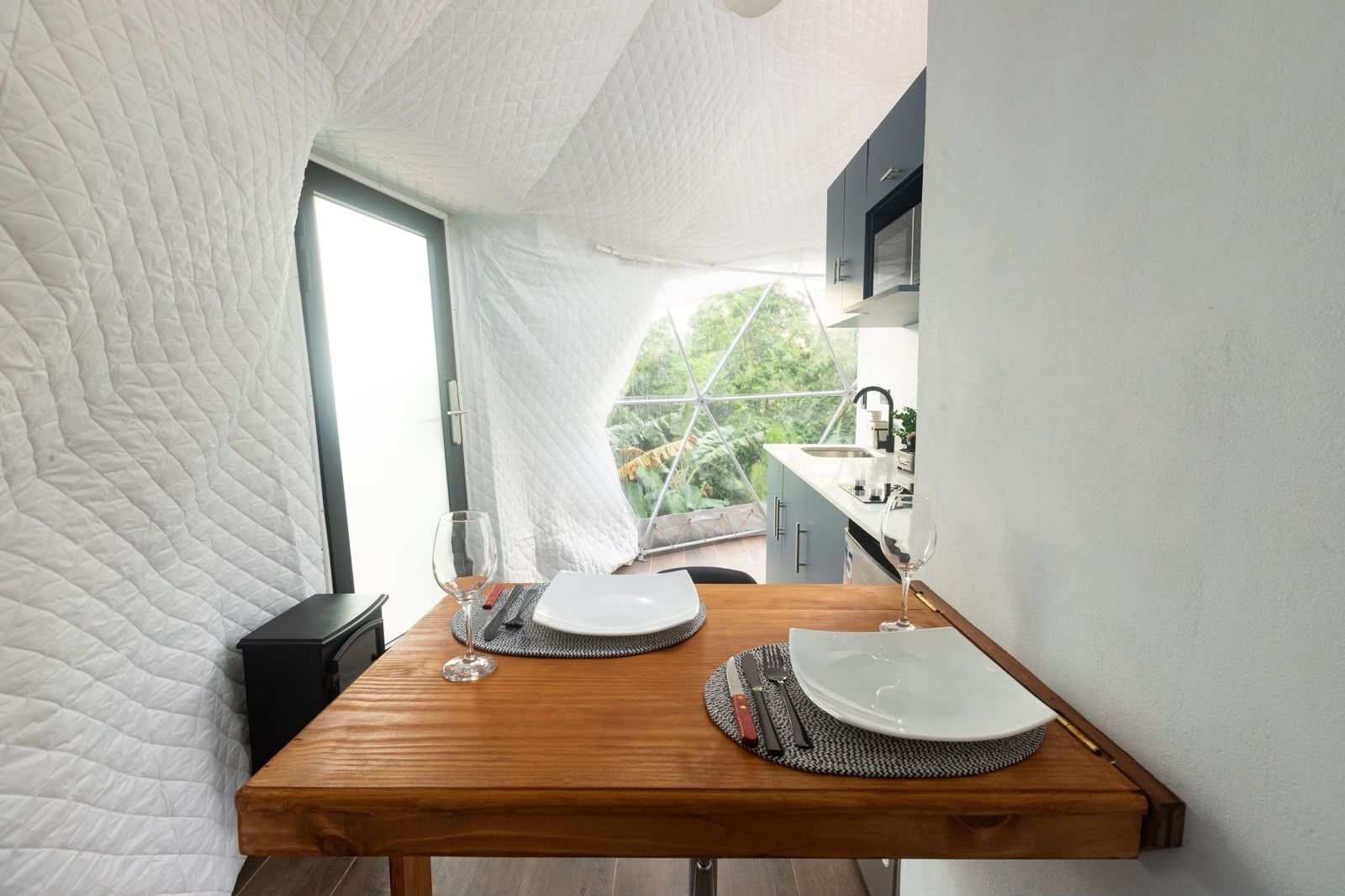 Una mesa de comedor dispuesta con platos, cubiertos y vasos, con una cocina a la vista al fondo.