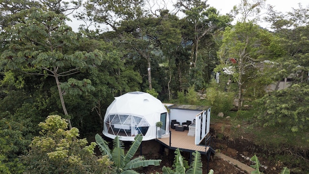 Une structure blanche en forme de dôme, surélevée, dotée d’une terrasse en bois et d’un espace pour s’asseoir, le tout entouré d’une végétation luxuriante.