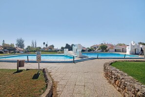A swimming pool with a stone pathway leading to it, surrounded by white buildings and greenery.