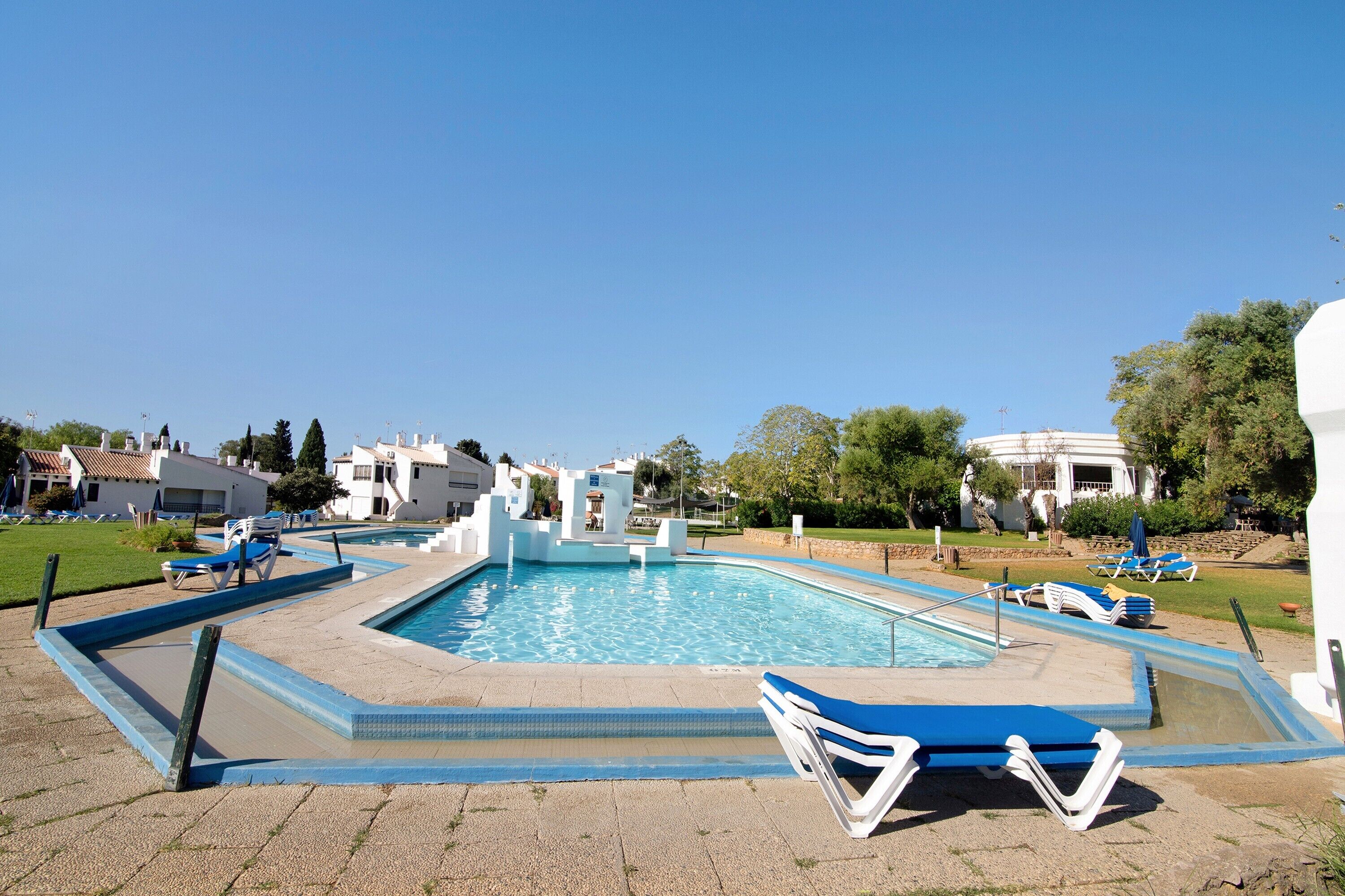 Ein Swimmingpool mit blauen und weißen Liegestühlen, einem wolkenlosen Himmel und umgebenden Gebäuden.