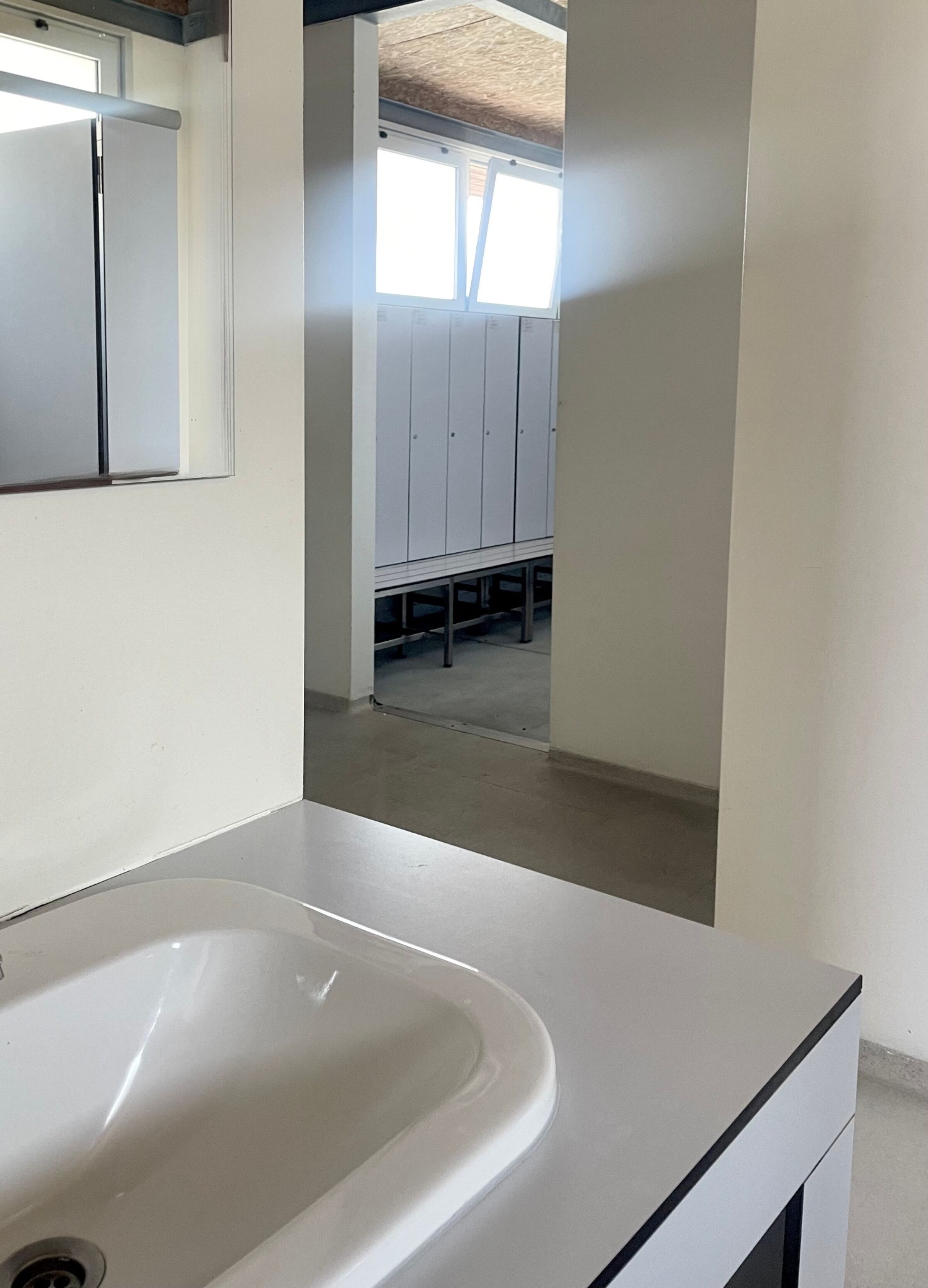 Une salle de bain avec un lavabo blanc, un miroir et une vue sur un vestiaire, visible à travers une porte.